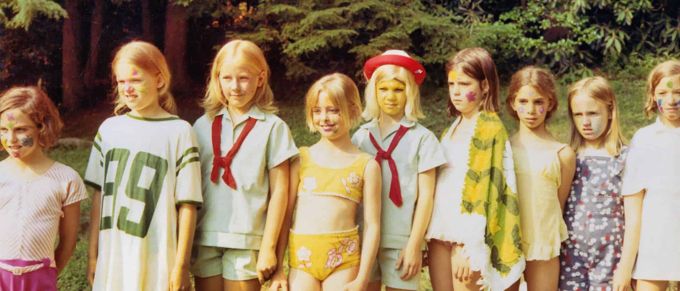 A group of children standing outdoors with colorful face paint, some wearing camp uniforms and others in casual or swimwear.