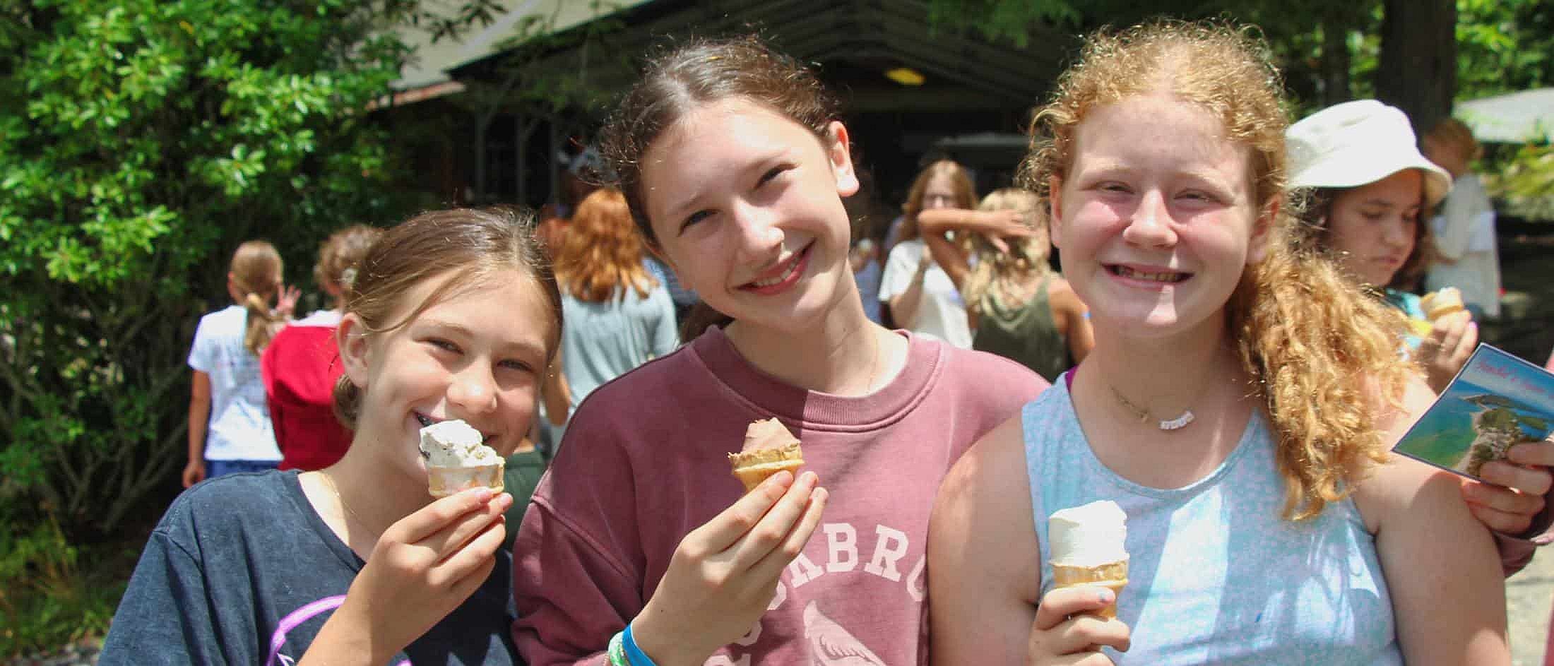 Three girls outdoors holding and eating ice cream cones, smiling toward the camera on a sunny day.