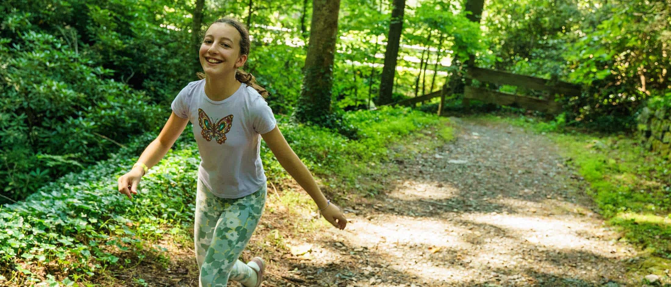 A smiling girl in a butterfly shirt and patterned leggings walks along a sunlit dirt path surrounded by green woods.