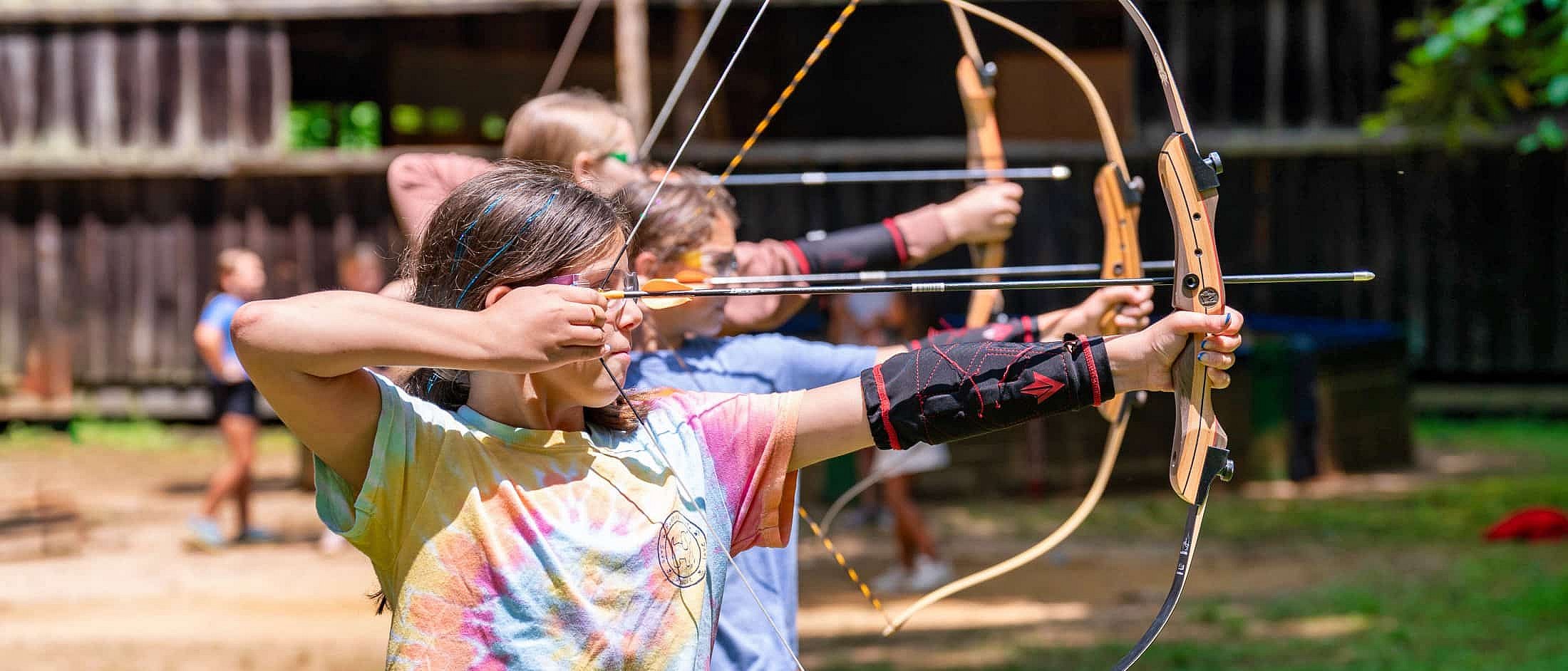 Three campers stand side by side outdoors, drawing bows and aiming arrows during an archery lesson.