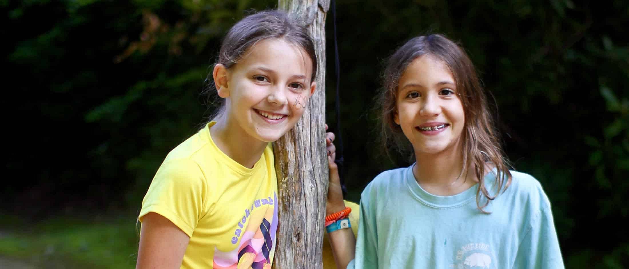 Two children smiling while standing outdoors beside a weathered wooden post with trees in the background.