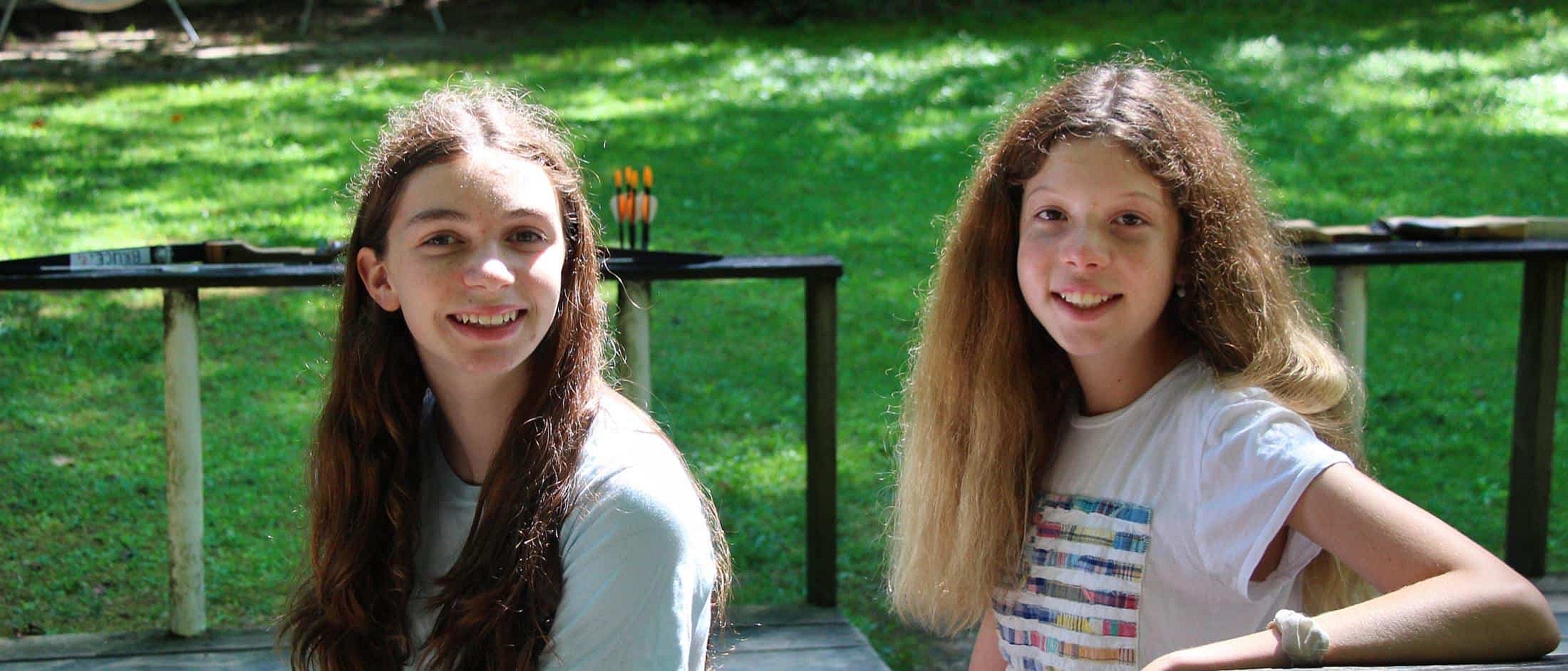 Two girls with long hair smile while sitting outside on a bench with archery targets in the background.