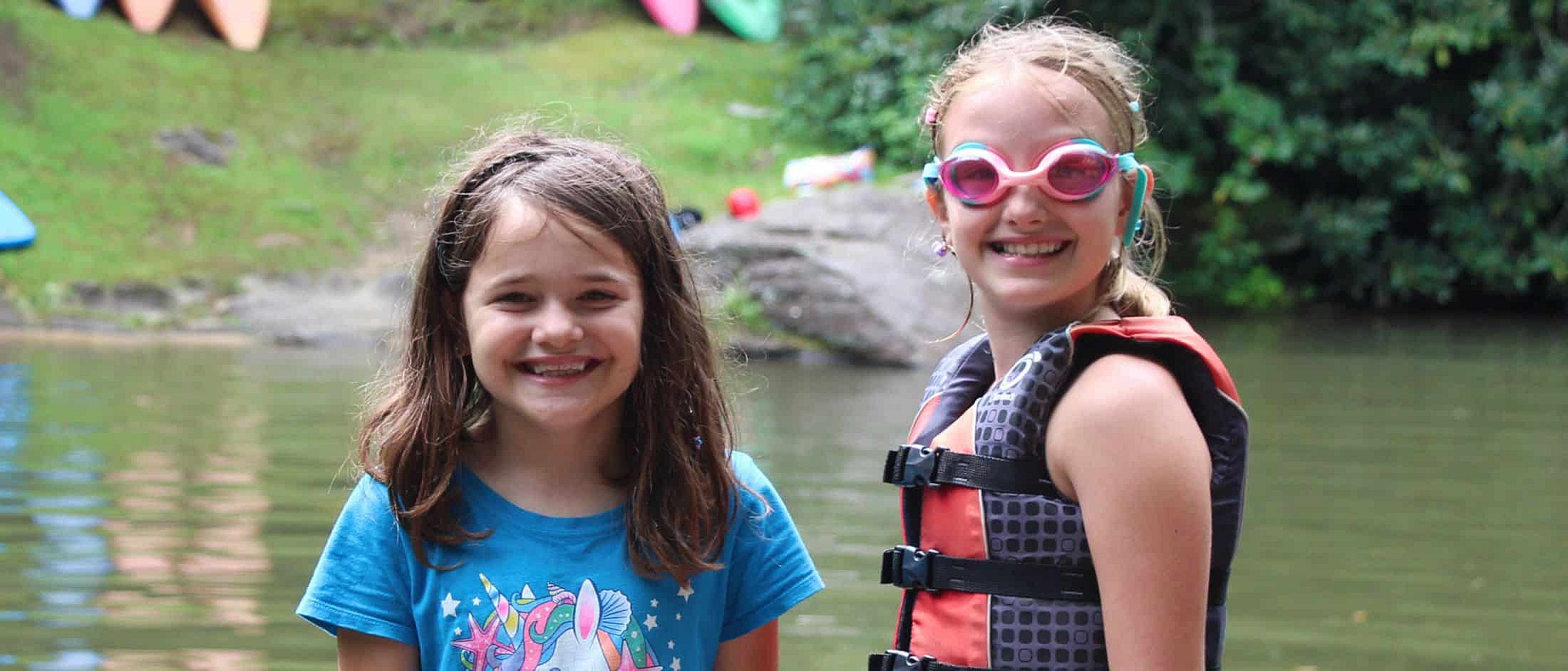 Two children smiling by a lake, one wearing pink swim goggles and a life jacket, the other in a blue unicorn t-shirt.