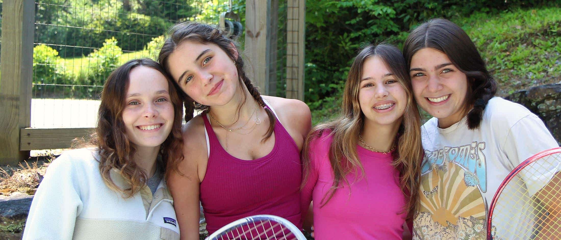 Four girls smiling and posing together outdoors, two holding tennis rackets, with a wooden fence and greenery in the background.