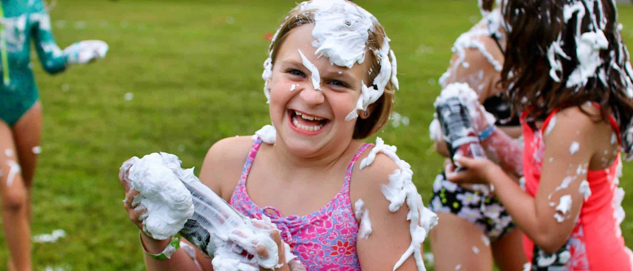A child laughing with foam covering their hair, face, and arms while holding a foam-filled bottle outdoors on grass.