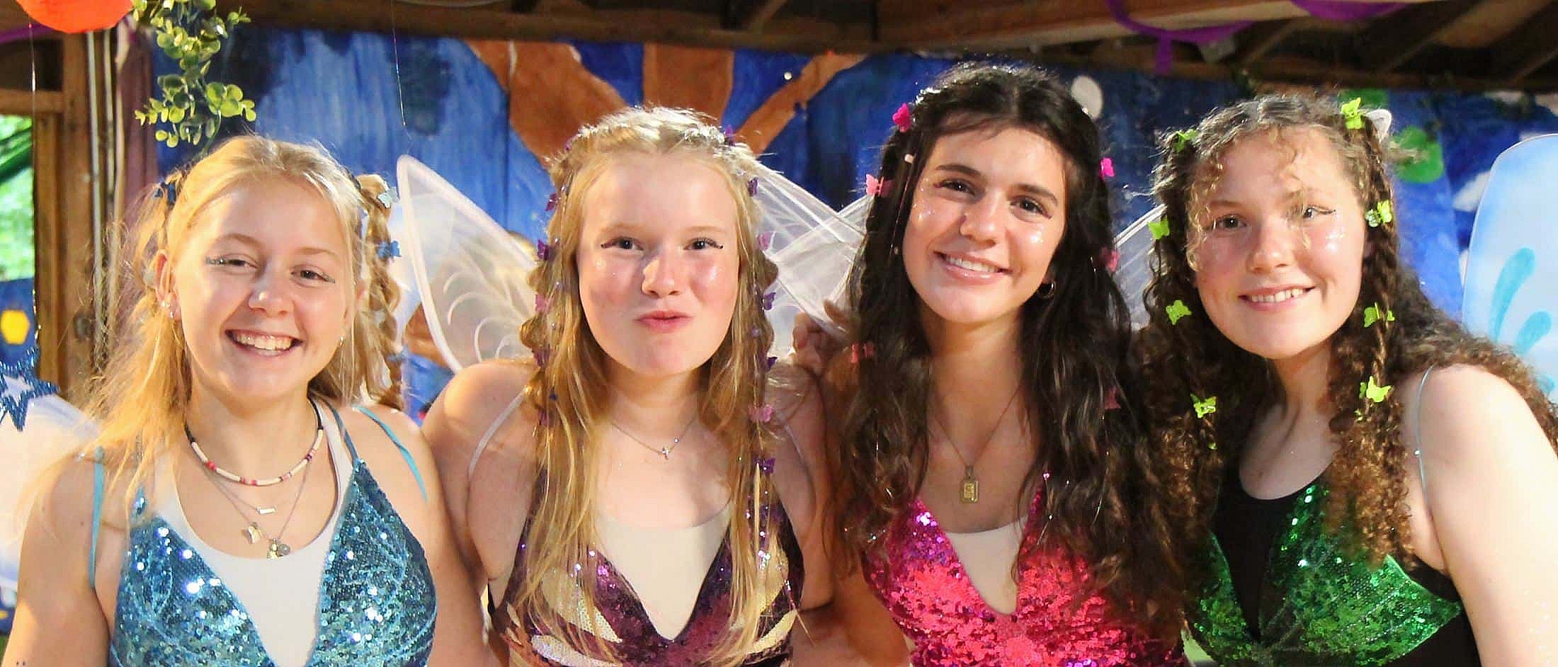 Four teens wearing colorful sequined fairy costumes with wings and decorated hair smile indoors against a painted backdrop.