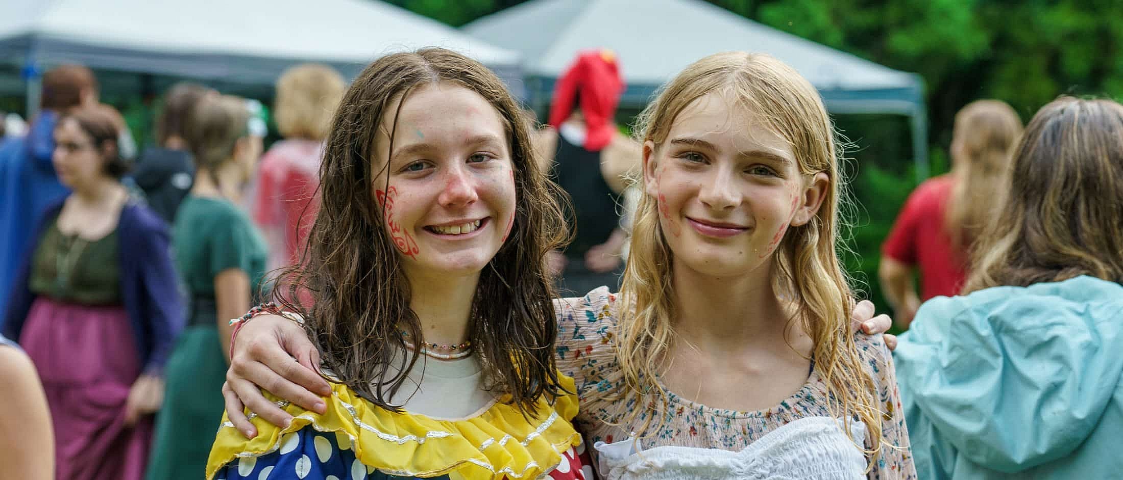 Two wet campers with face paint smiles with their arms around each other at an outdoor event with tents and people in the background.