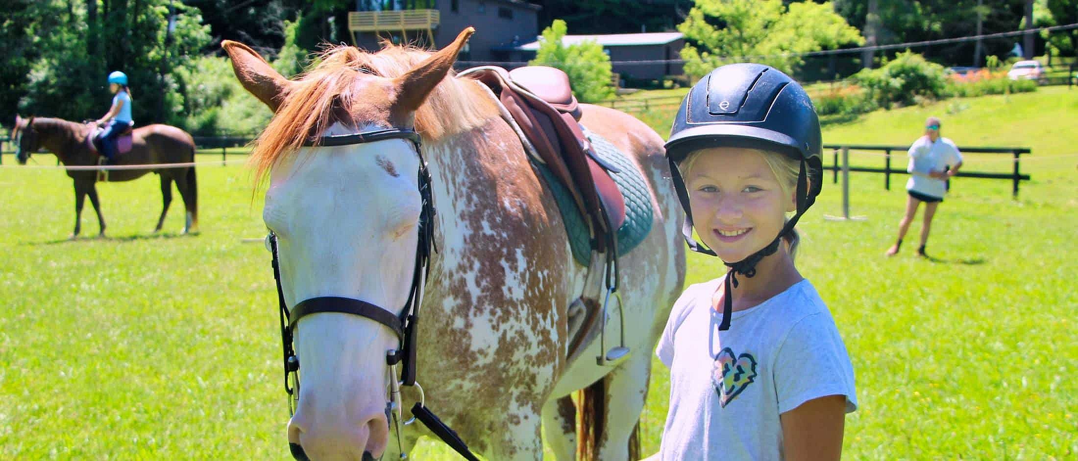 A child wearing a riding helmet smiles next to a saddled pinto horse in a sunny grassy field with other riders and an instructor in the background.