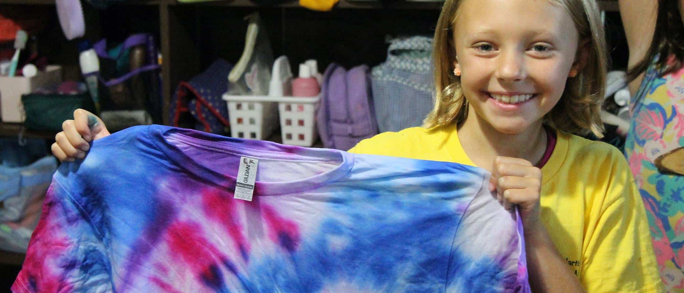 Smiling camper holds up a colorful tie-dyed shirt with swirling blue, purple, and pink patterns.