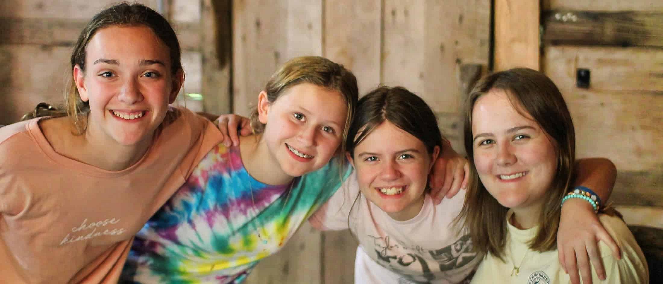 Four smiling girls sit close together indoors with their arms around each other’s shoulders.