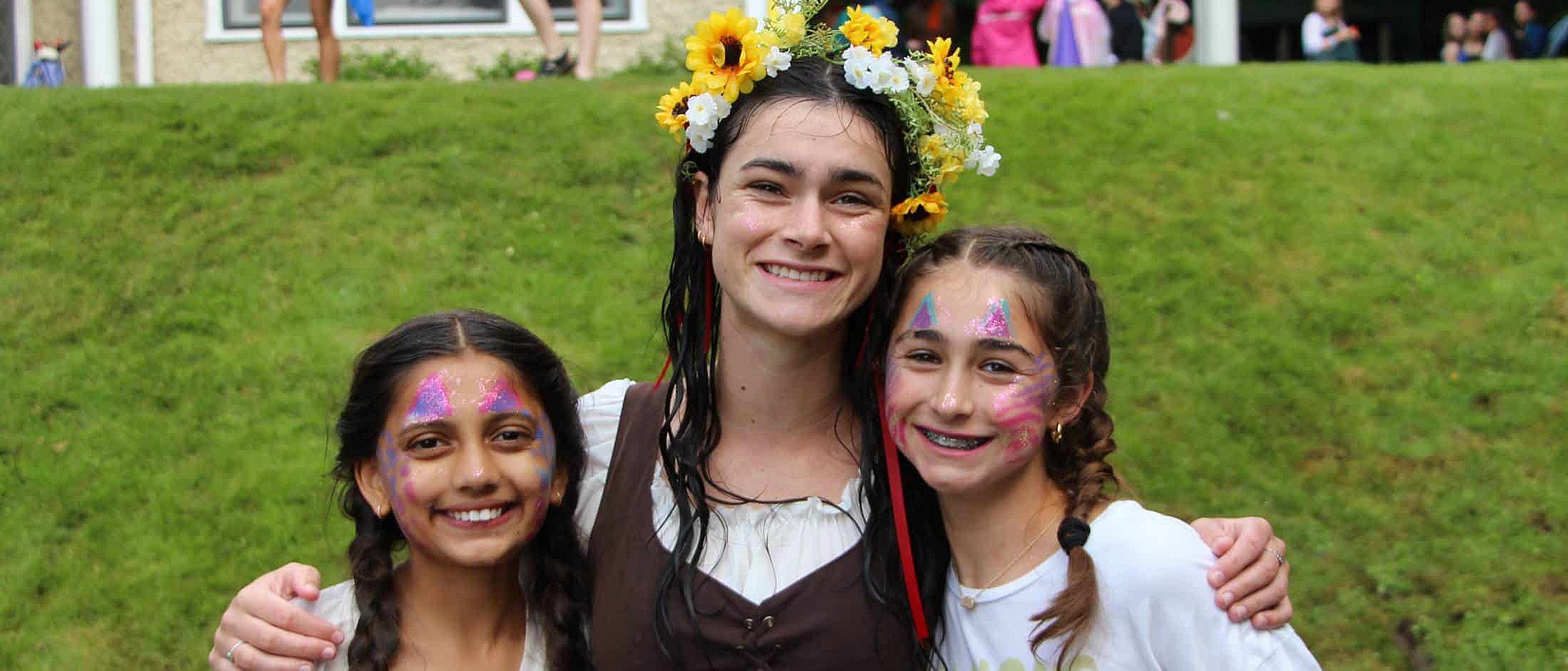 Three girls with face paint smile together outdoors, with one wearing a flower crown and their arms around each other.