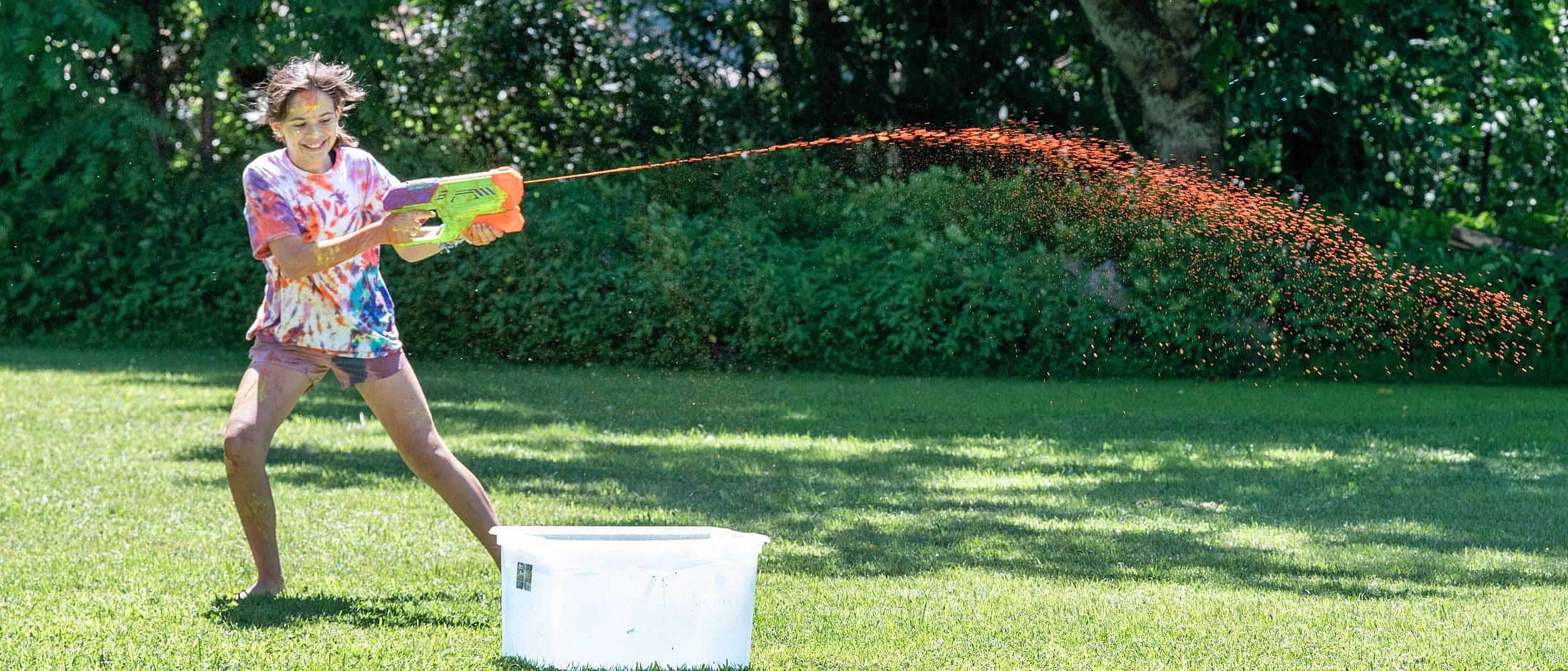 A person in a tie-dye shirt sprays orange paint from a water gun across a grassy yard near a white plastic tub.