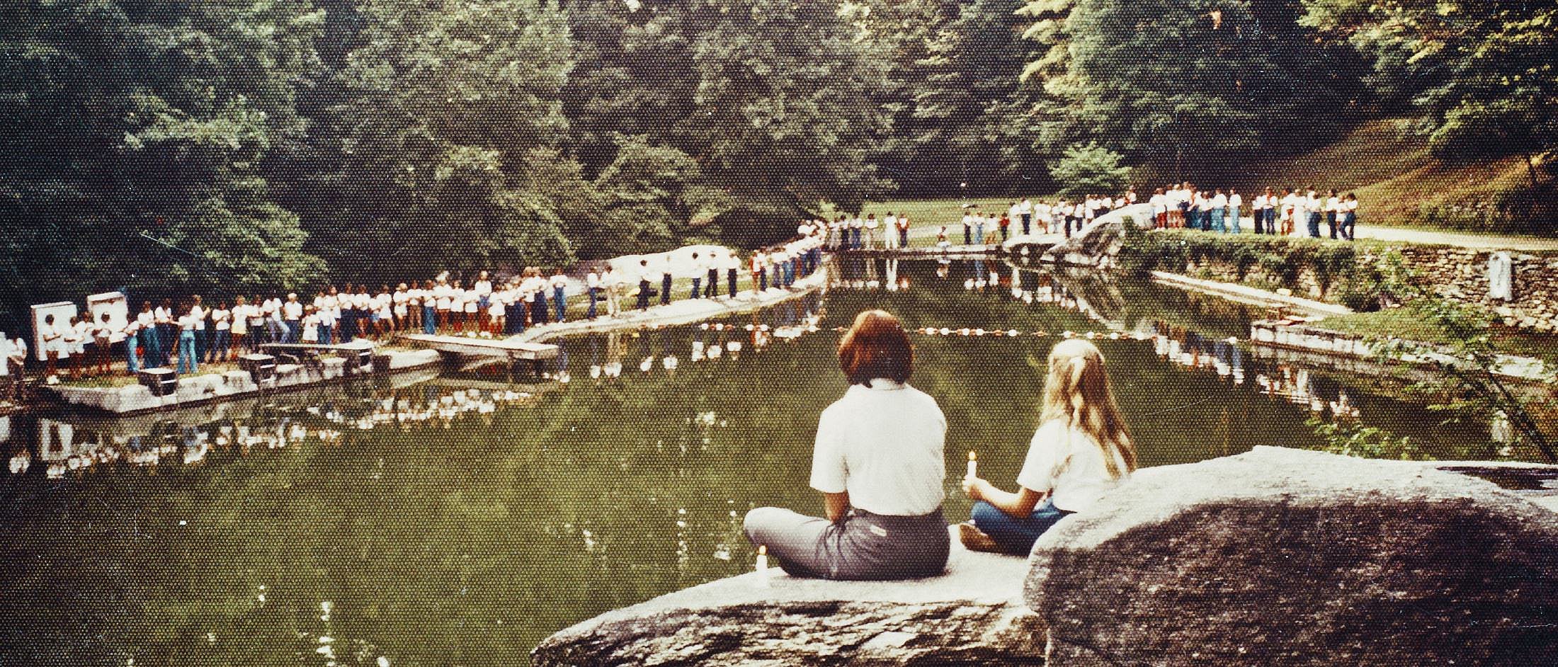 Two people sit on a large rock by a calm lake while a long line of people stands along the opposite shore surrounded by trees.