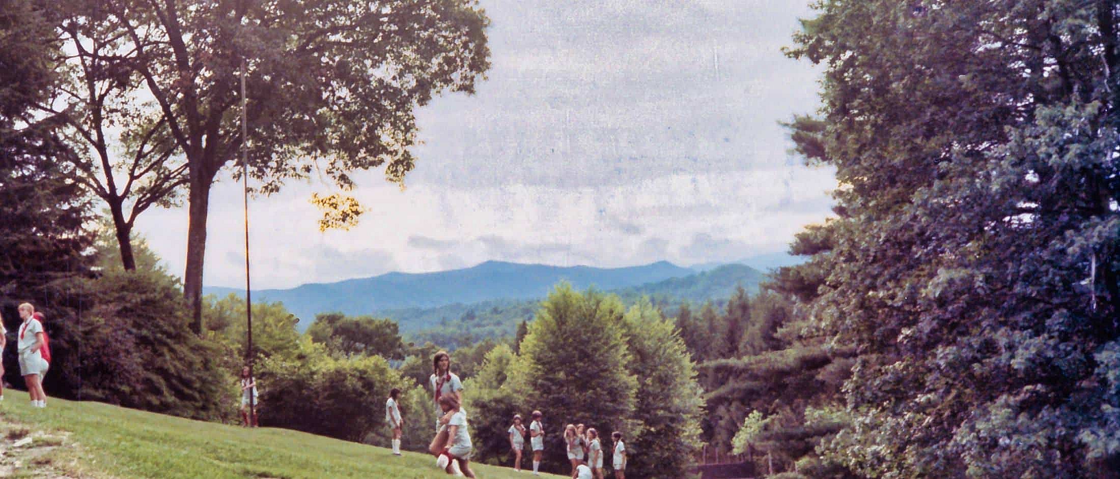 Campers stand and play on a grassy hill surrounded by trees with mountain ranges visible in the distance under a cloudy sky.
