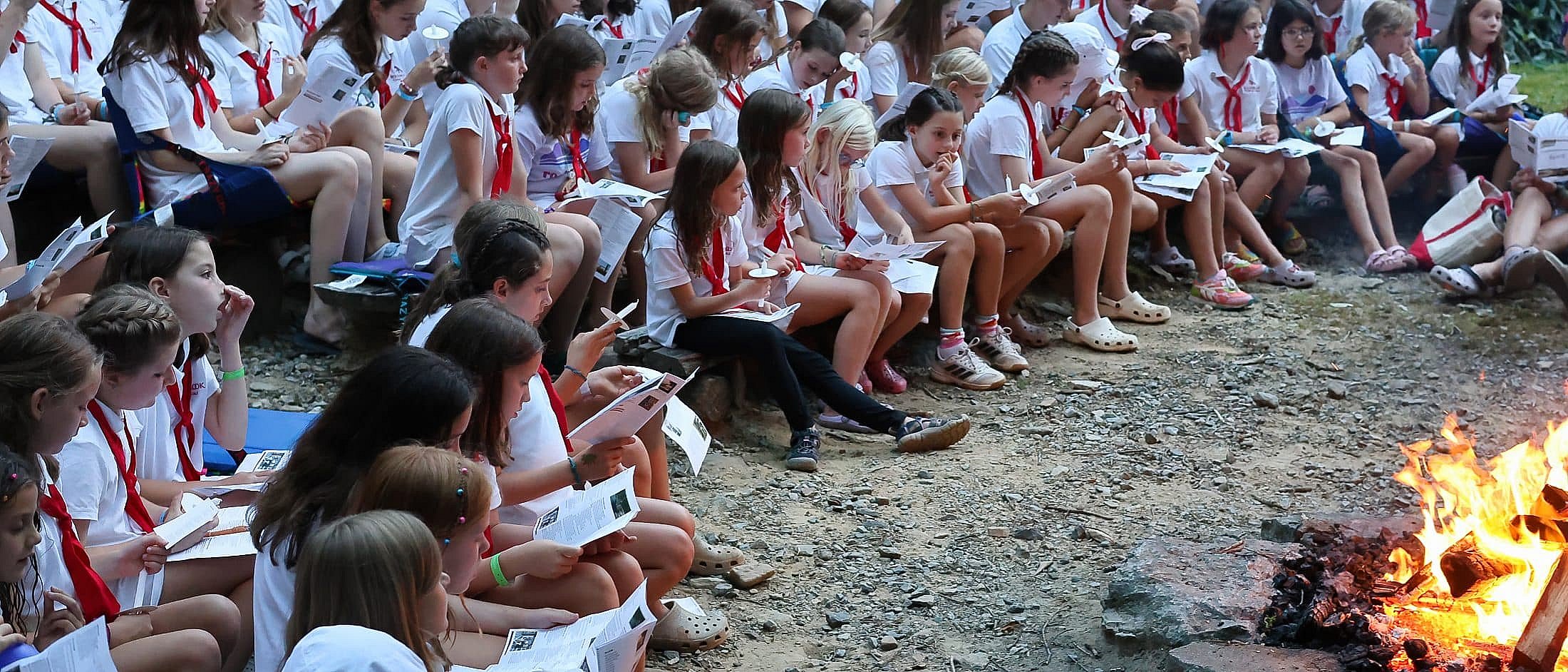 Campers sitting closely around a campfire, holding papers and candles during a group ceremony outdoors.