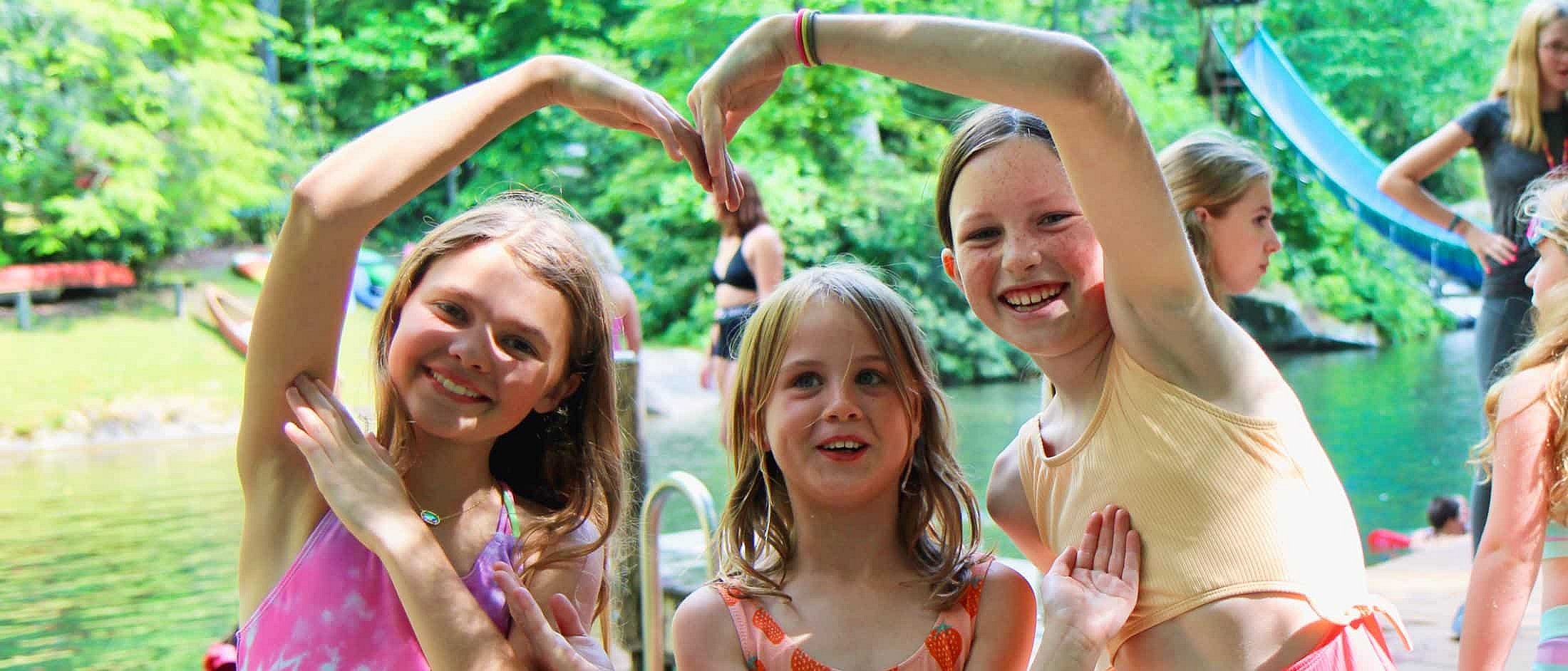 Three campers smile by a lake as two form a heart shape with their arms over the third camper’s head.
