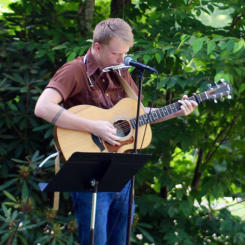 Clint Roberts plays an acoustic guitar and harmonica simultaneously while singing into a microphone outdoors with greenery in the background.