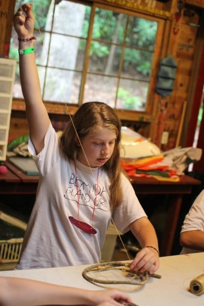 A camper carefully weaves string through a circular frame while sitting at a table in a rustic craft room.