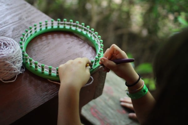 Hands weaving yarn on a green circular loom placed on a wooden surface outdoors.