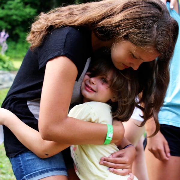 A person with long hair leans down to hug a smaller child who is smiling and holding on tightly outdoors.