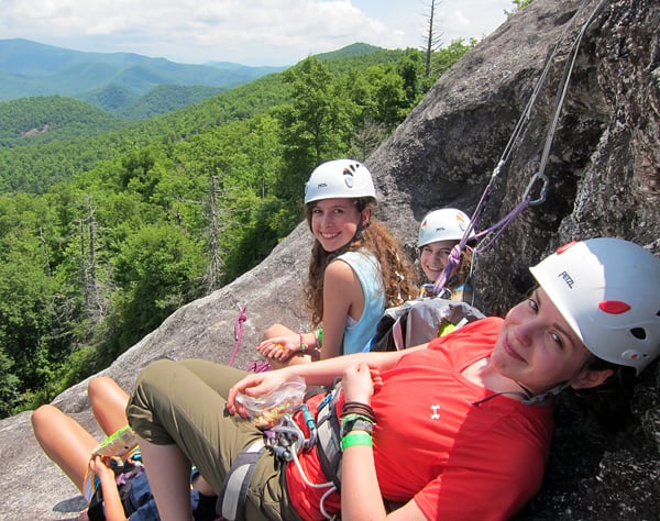 Three climbers in helmets and harnesses rest on a rock ledge secured by ropes with forested mountains in the background.