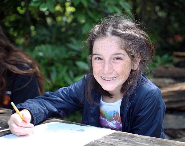 A camper with braided hair smiles while drawing with a pencil at an outdoor wooden table surrounded by greenery.