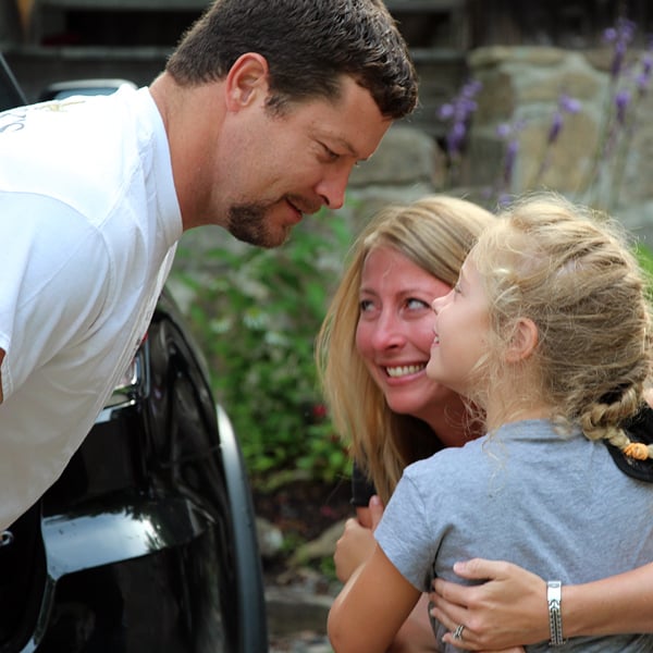 A father leans down smiling as a mother embraces a young girl campers outdoors near a vehicle being dropped off at summer camp.