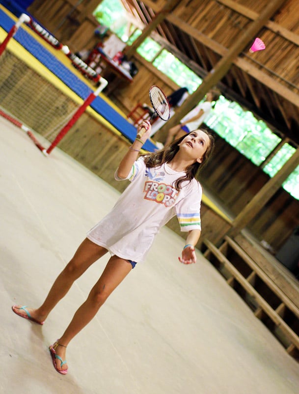 A camper in a large indoor space prepares to hit a pink shuttlecock with a badminton racket.